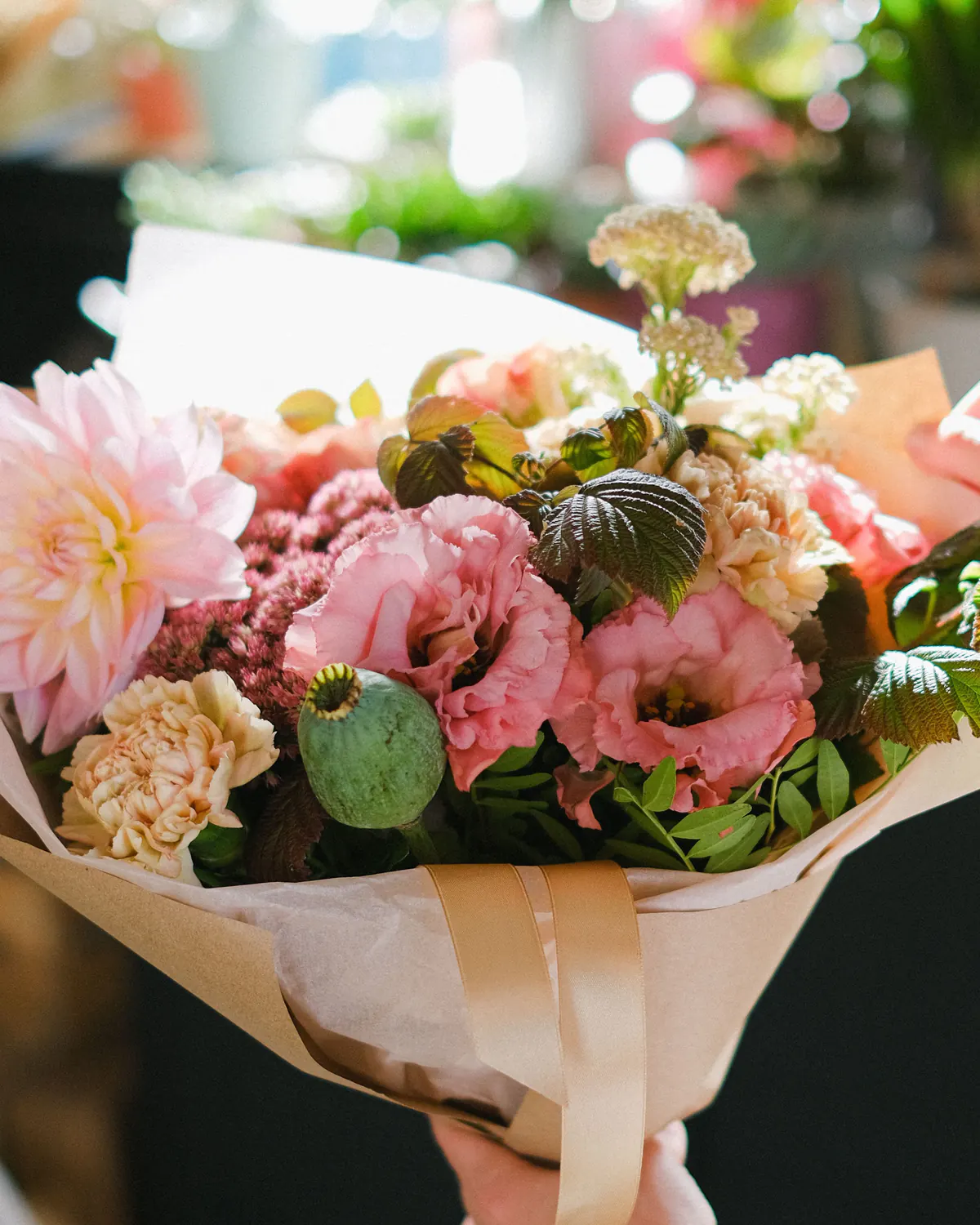 A large hand-tied bouquet of pink lisianthus, dahlia and carnations wrapped in kraft paper with a satin ribbon.