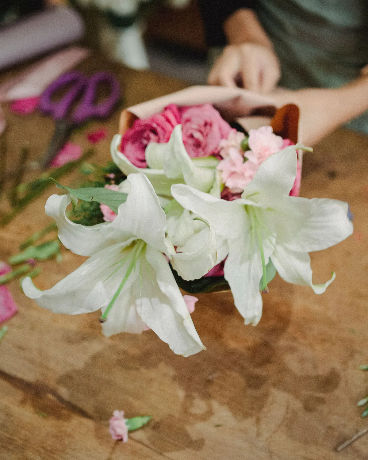 A small bouquet of white lilies and pink roses being wrapped in kraft paper on a florist's table.